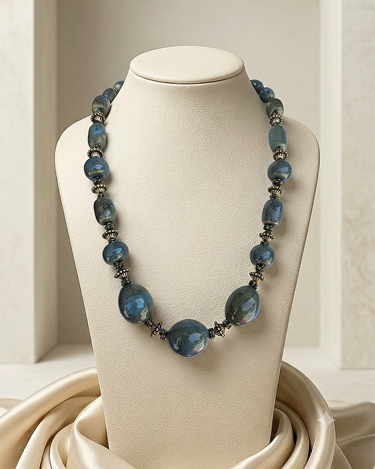 A beaded necklace with faceted blue beads and dark metallic spacer accents, displayed on a cream jewelry bust with draped satin fabric at the base.
