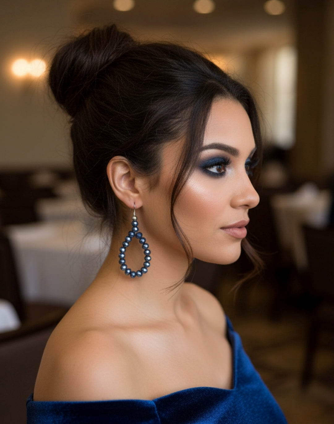 Woman wearing a blue off-shoulder top and large earrings with a blurred indoor background