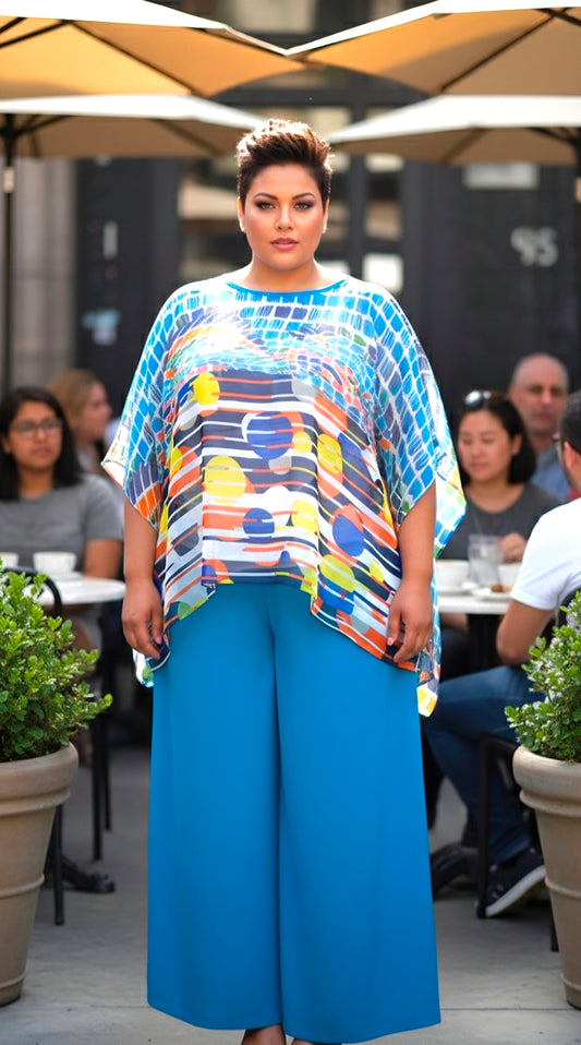 Woman wearing a colorful top and blue pants standing outdoors with people and plants in the background.