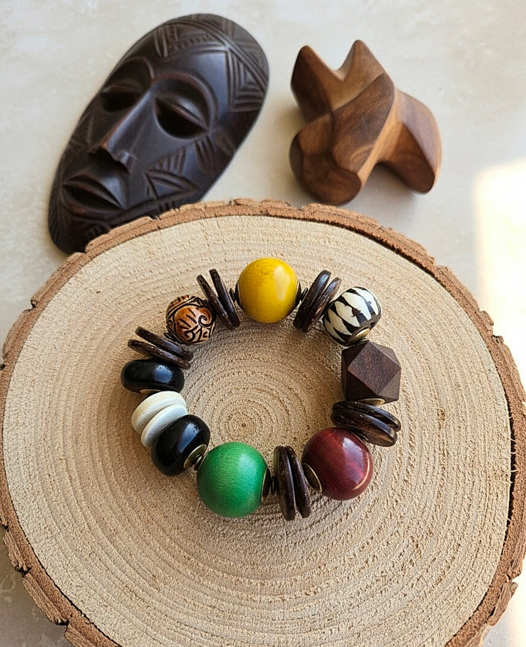 Colorful beaded bracelet on a wooden surface with a dark mask and wooden figure in the background.