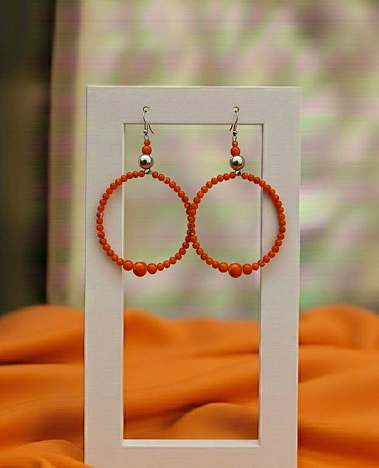 Pair of orange hoop earrings with silver beads on a white card against a blurred background.