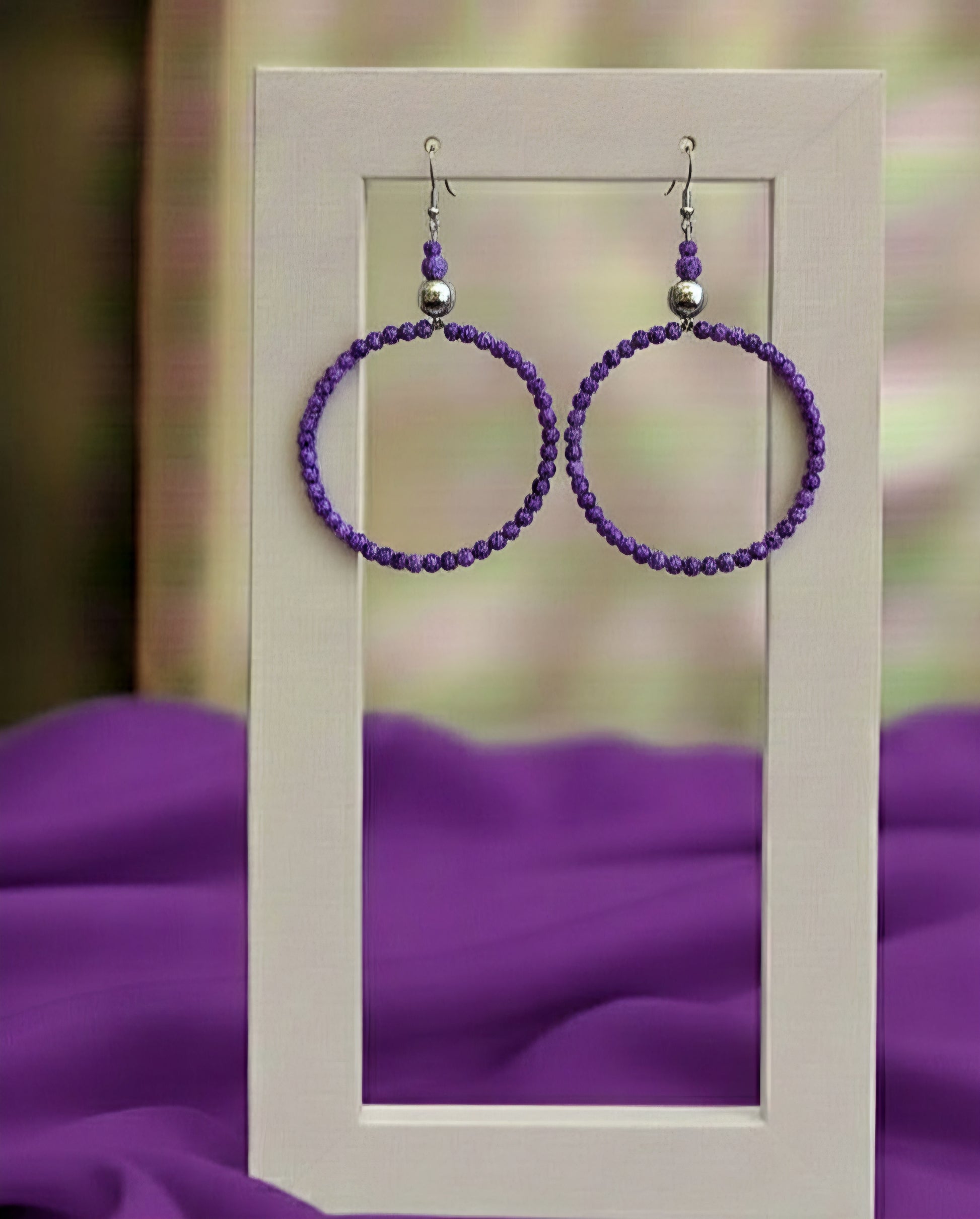 Purple beaded hoop earrings on a white card with a blurred purple fabric background