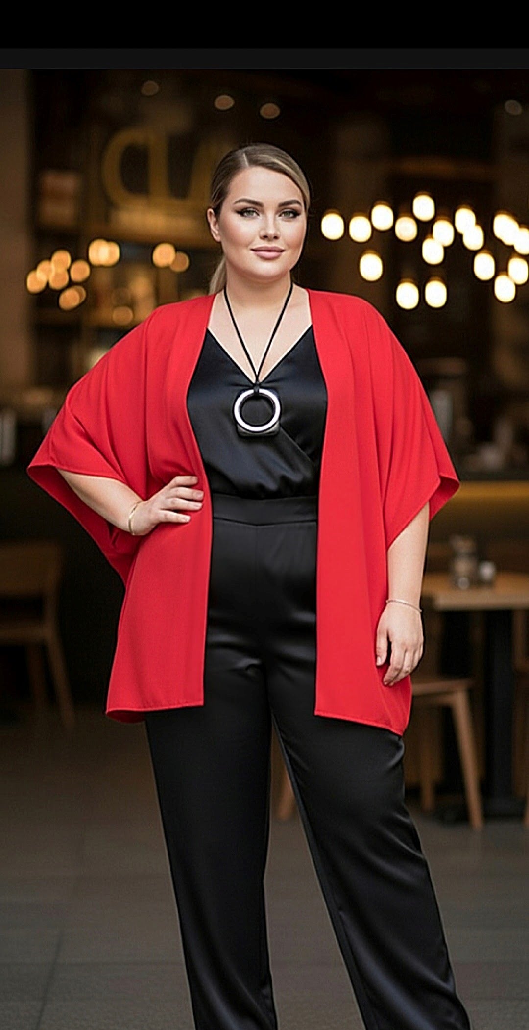 A woman modeling a bright red open-front cape-style jacket over a black satin jumpsuit, standing in a softly blurred indoor setting with warm lights.