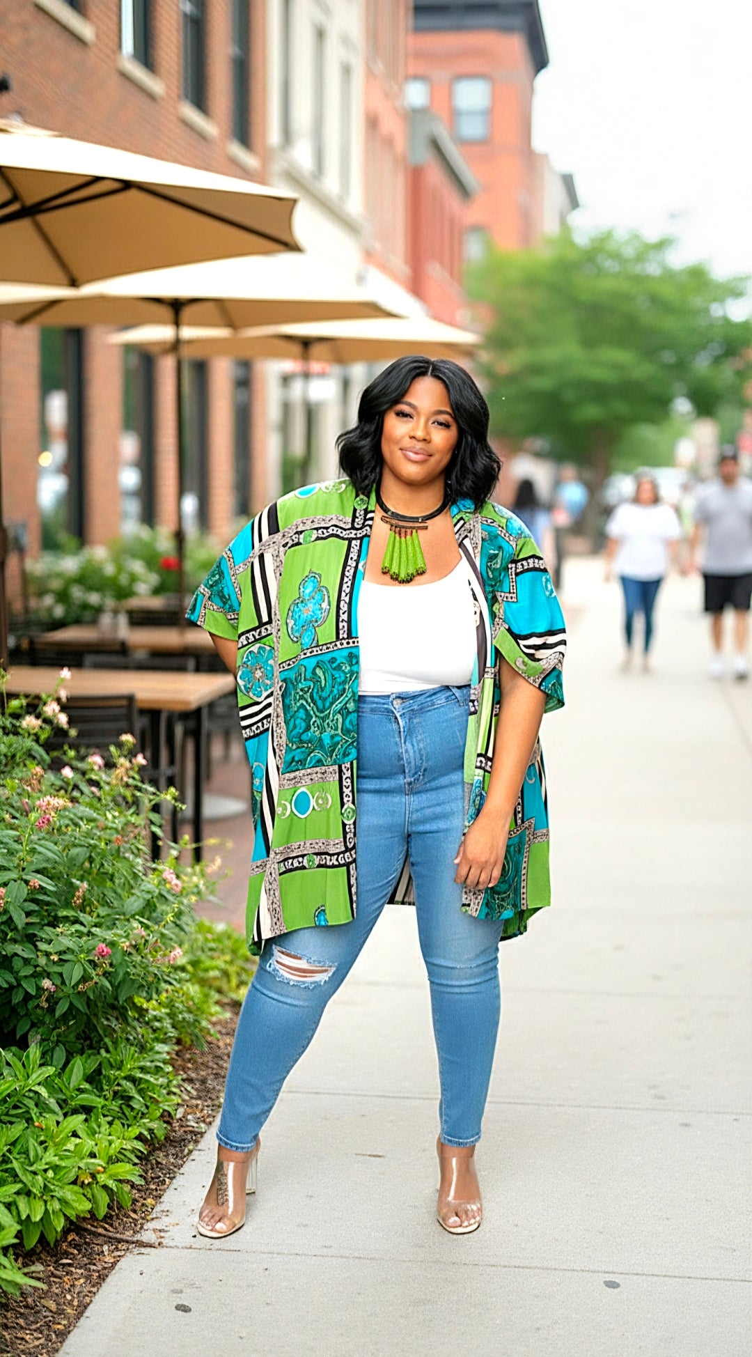 Woman in colorful jacket and blue jeans standing on a sidewalk with buildings and people in the background