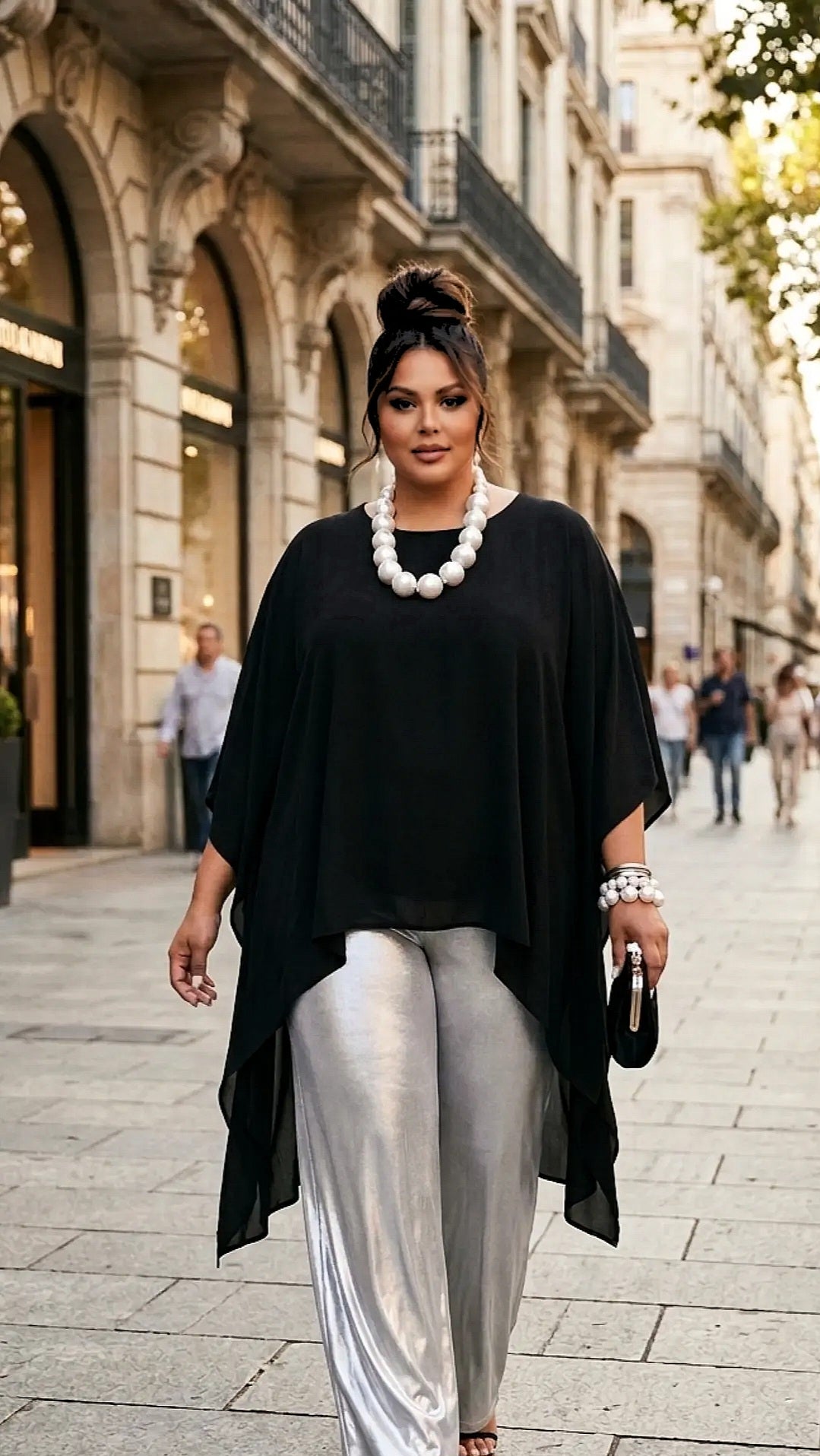 A woman walking down a city street wearing a black draped poncho top, shiny silver wide-leg pants, a chunky white pearl necklace, and holding a black clutch.