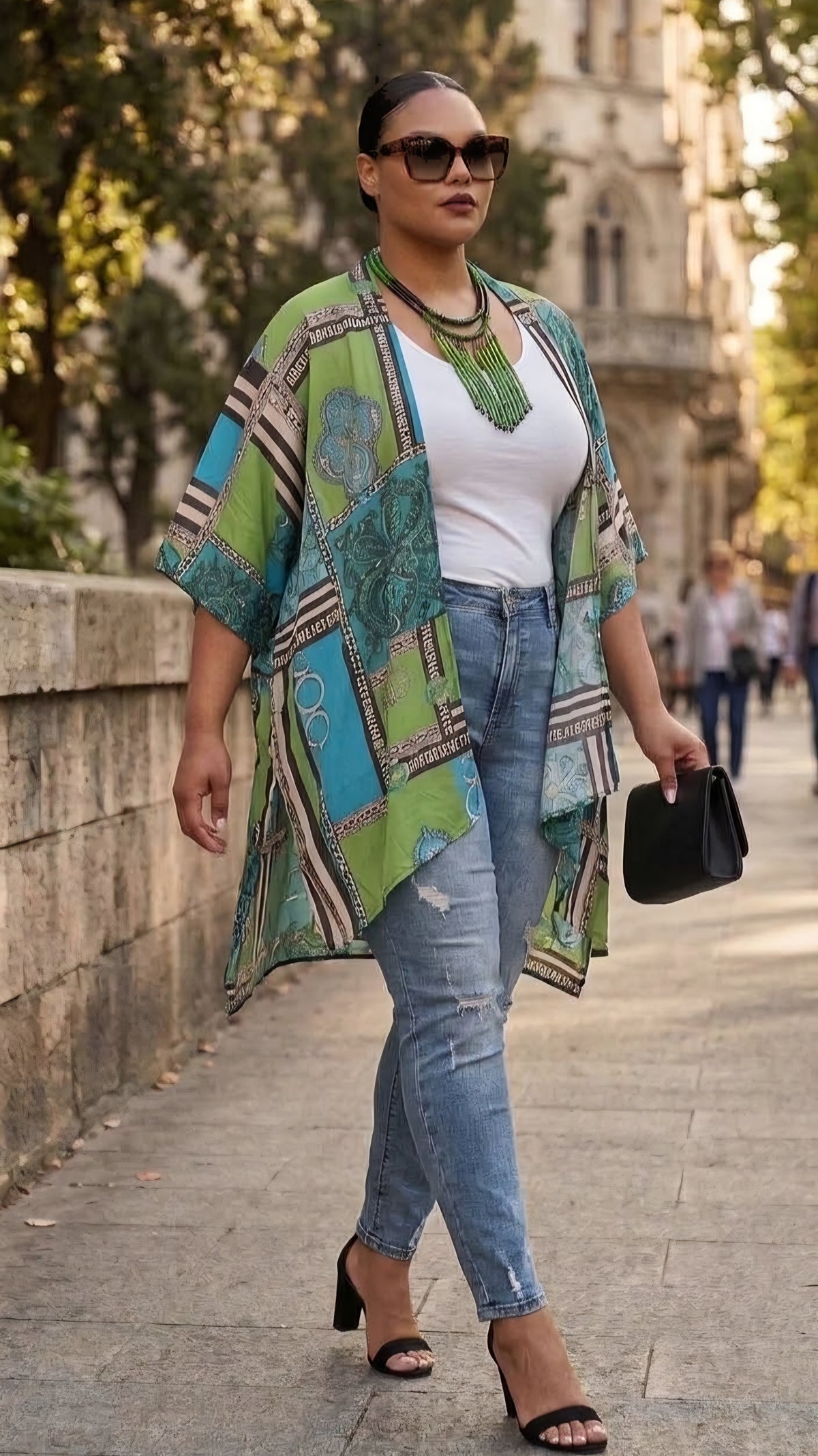 A woman walking outdoors on a city sidewalk wearing a green and blue patterned open-front cardigan over a white top and light-wash jeans, with black heeled sandals and a small black handbag.