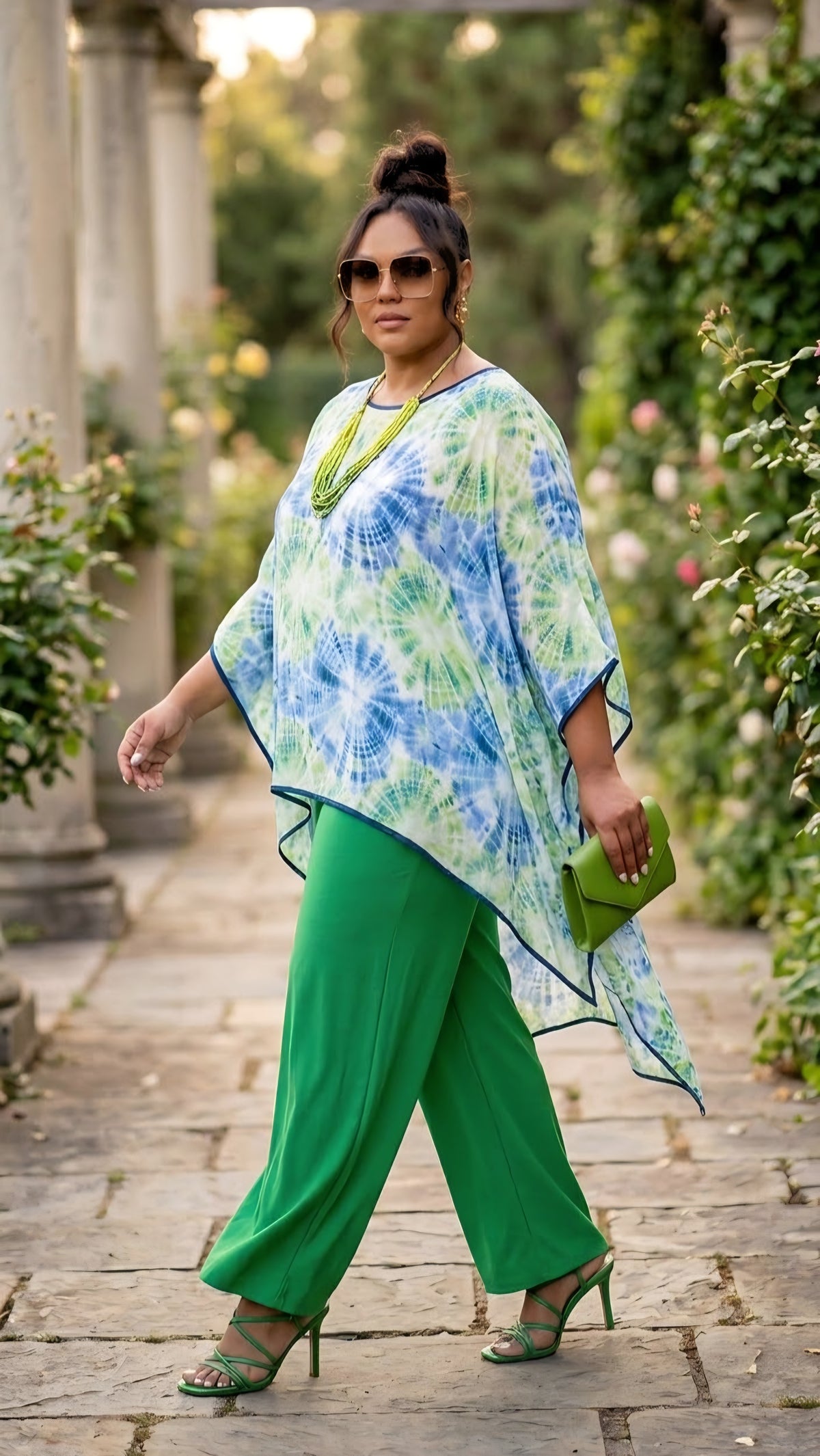 A woman walking outdoors on a stone path wearing a green and blue tie-dye poncho top, bright green wide-leg pants, and green heels, holding a small green clutch.