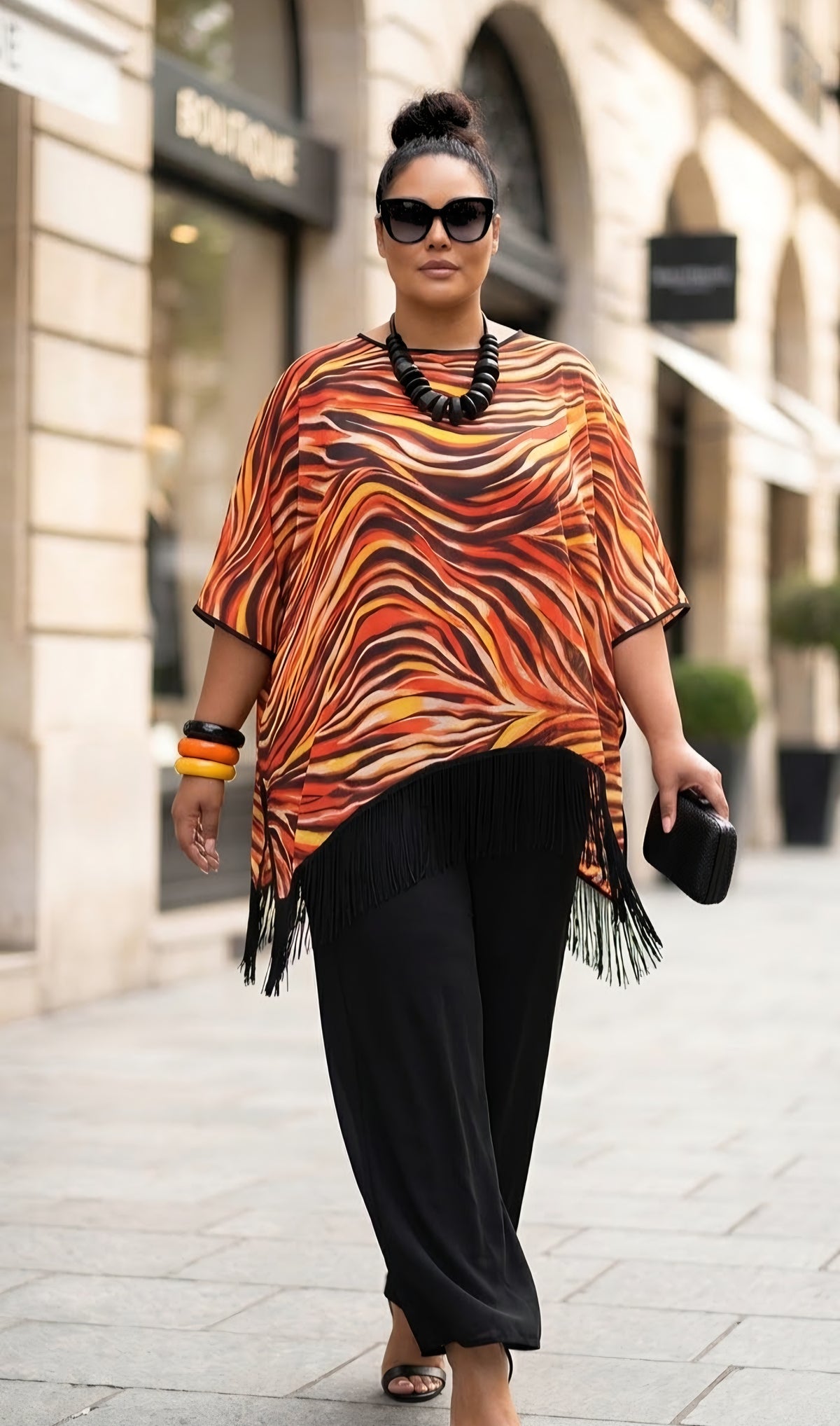A woman walking down a city street wearing a vibrant orange, brown, and black wavy patterned tunic top with fringe trim, paired with black pants.