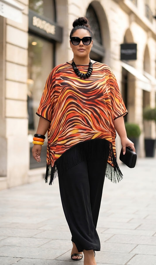 A woman walking down a city street wearing a vibrant orange, brown, and black wavy patterned tunic top with fringe trim, paired with black pants.