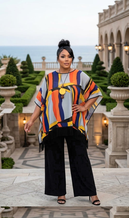 A woman stands on a stone terrace in front of a grand building and the ocean, wearing a loose short-sleeve top with bold vertical stripes in orange, yellow, blue, white, and black, paired with black wide-leg pants.