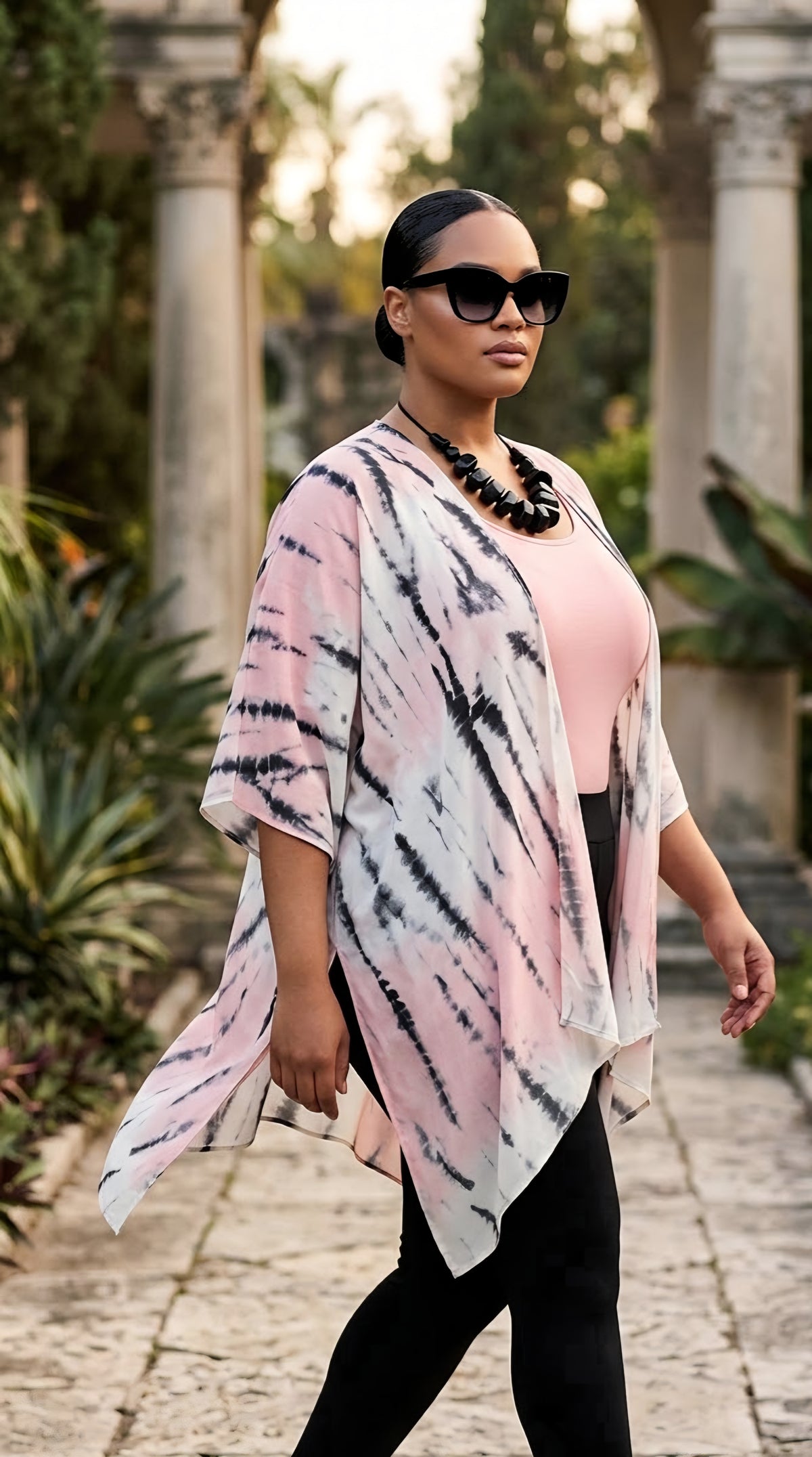 A woman walking outdoors in a garden courtyard wearing a pink, black, and white tie-dye kimono cardigan over a pink top and black pants.