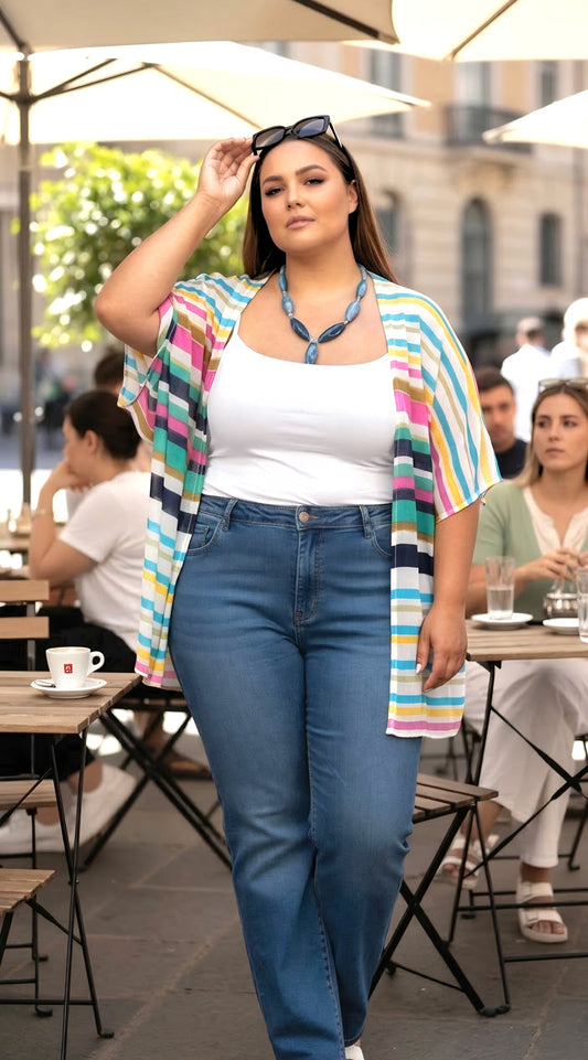 A woman walking outdoors at a cafe wearing a white tank top, blue jeans, a colorful striped open-front cardigan, and a blue necklace.
